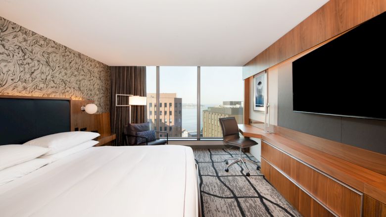 Modern hotel room with a large bed, wall-mounted TV, desk, chair, floor-to-ceiling window, and city view.
