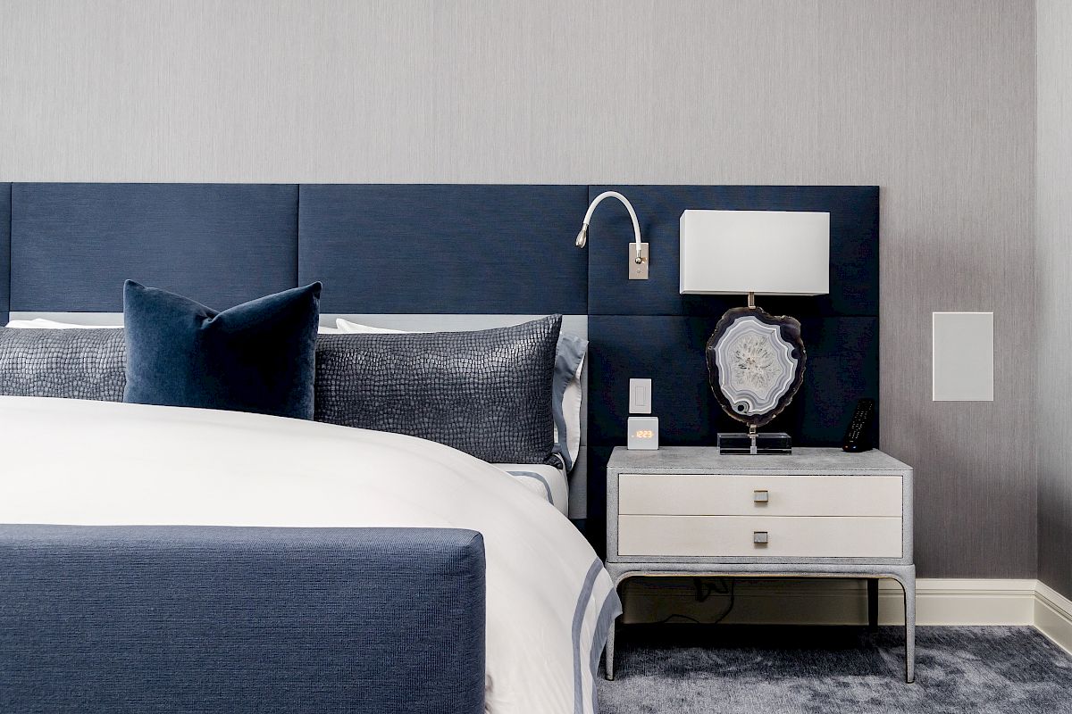 A modern bedroom featuring a blue upholstered bed, matching pillows, a white nightstand with a lamp, and a blue and gray color scheme.