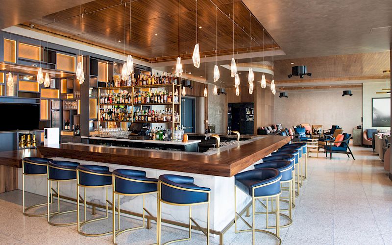 A modern bar with a wooden counter, overhead pendant lights, and blue barstools. Shelves stocked with bottles, lounge seating in the background.