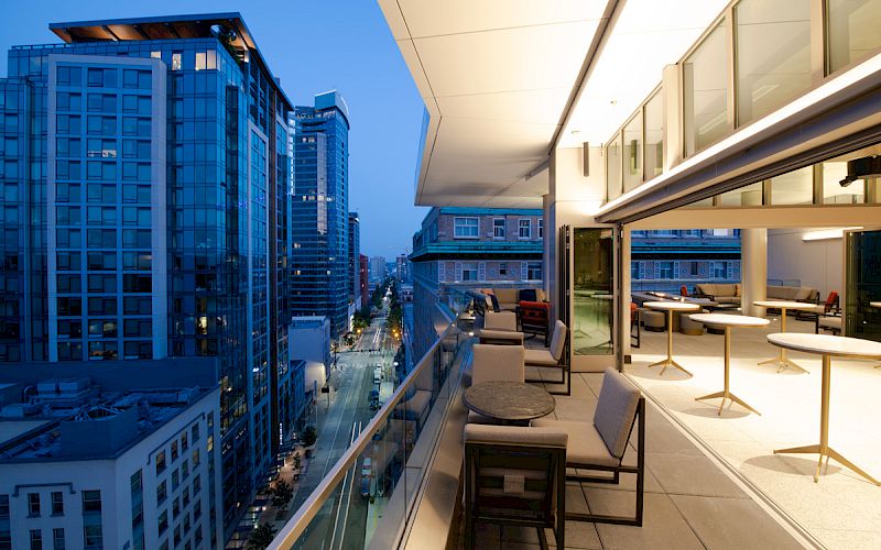 The image shows a modern outdoor balcony with seating, tables, and a view of city buildings. The sky appears to be at dusk or early evening.