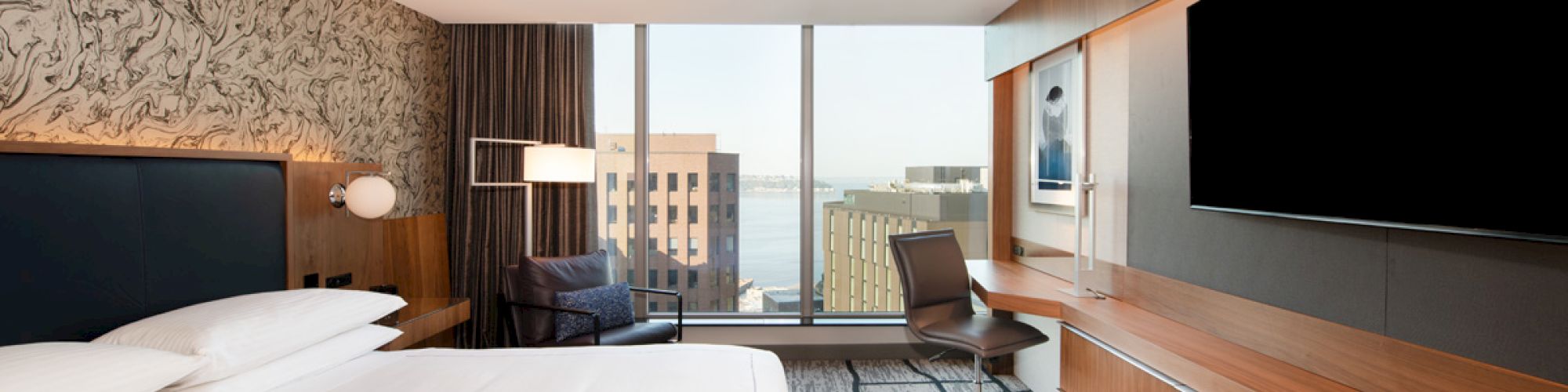A modern hotel room features a large bed, work desk, chair, and flat-screen TV with a cityscape view from a floor-to-ceiling window.